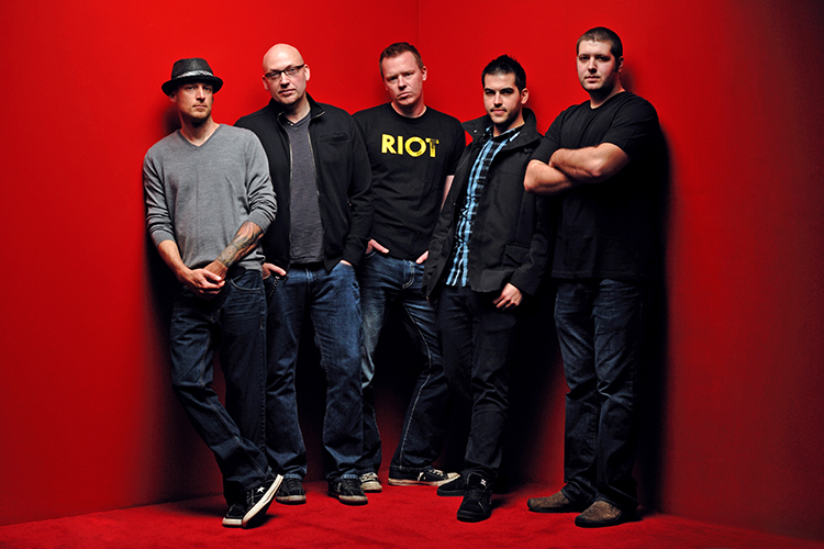 Five members of a musical band calmly stand together in a red room. They are all dressed in dark colors and are looking intently at the camera.
