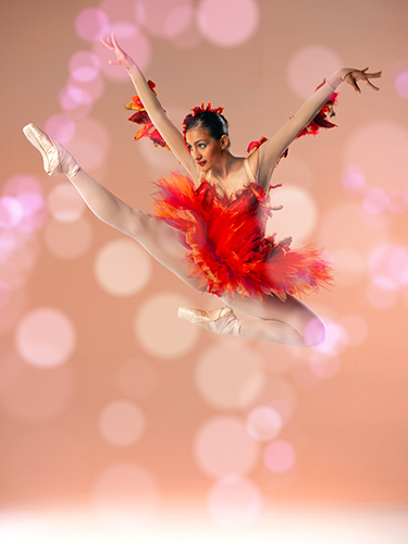 Ballet dancer in vibrant red and orange feathered costume poses gracefully leaps in the air against a soft, glowing pink background.