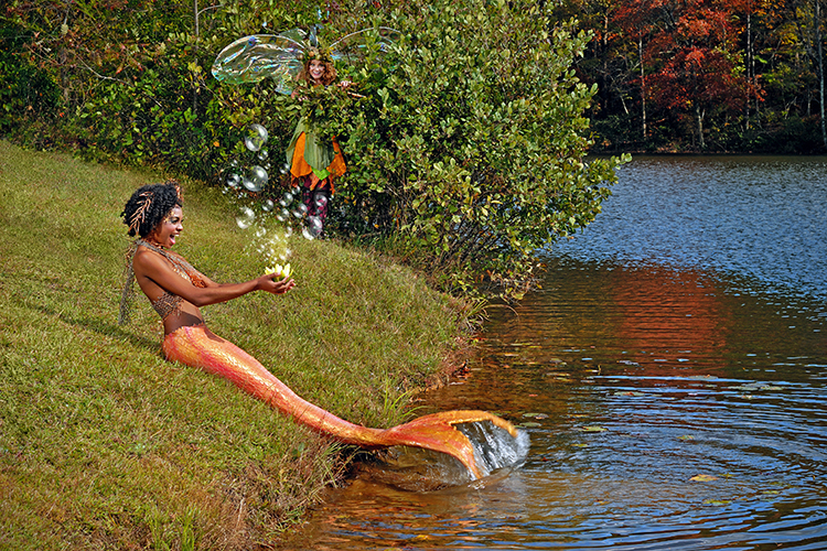 A mermaid with a vibrant orange tail sits by a lake, interacting with glowing bubbles. A fairy with translucent wings hides in the bushes nearby watching the activity.