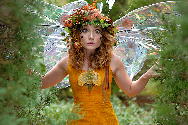 A woman with curly hair wears a vibrant fairy costume, featuring a floral headpiece and translucent wings. She stands amidst greenery, with a surprised expression on her face.