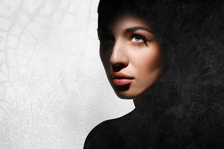 A woman with a contemplative expression is partially shrouded in shadow. The background features a textured, cracked surface merging with the darkness. The lighting highlights her eyes and lips.