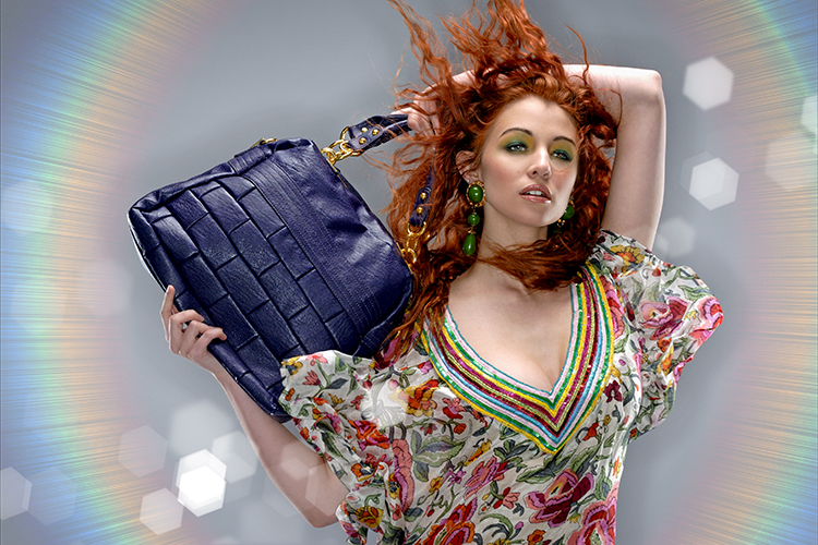 A woman with red hair poses confidently, holding a large blue handbag over her shoulder. She is wearing colorful earrings and a floral-patterned dress with a vibrant neckline, set against a backdrop with a circular light effect.