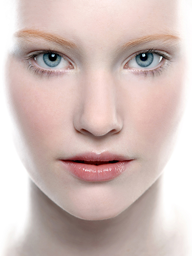 A close-up of a woman's face with fair skin, light blue eyes, and light eyebrows. The lighting is bright and the expression is neutral, with slight gloss on the lips.