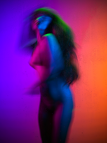 A blurred silhouette of a woman with long hair, standing against a multicolored background of blue, purple, and orange hues. Water droplets are visible on a surface in front of them, adding texture to the vibrant scene.