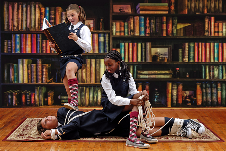 Three children in matching school uniforms are in a library with bookshelves. One child is reading a book, standing with a foot on a tied-up child lying on the floor. Another child is kneeling, holding rope with a playful expression.