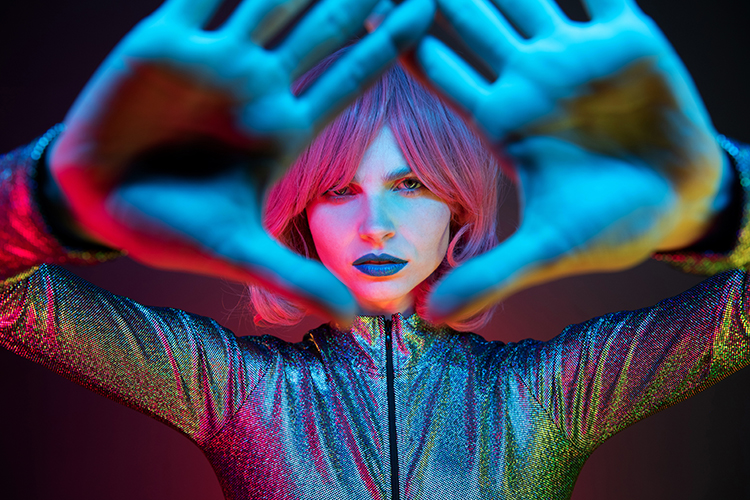 A woman with a pink wig and blue lipstick poses with hands framing their face, wearing a shimmering silver outfit. Bright, colorful lighting casts a mix of blue, pink, and red hues on them, creating a vibrant, dramatic effect.