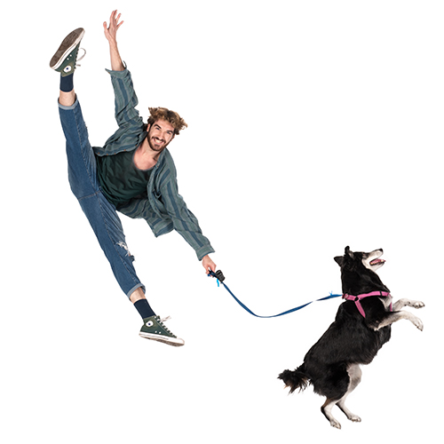 A man energetically leaps into the air, holding onto a leash with a playful black and white dog pulling enthusiastically. He wears jeans, a green shirt, and a striped jacket. The background is plain white.