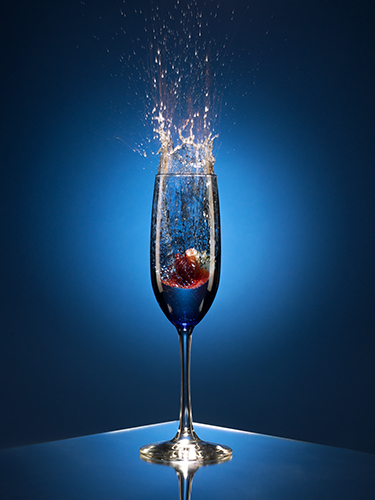 A splash erupts as a fruit is dropped into a clear champagne flute, filled with liquid, set against a blue background. Light reflects off the liquid, capturing the motion and energy of the splash.