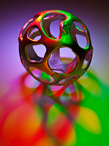 A spherical, openwork metal sculpture casts colorful shadows in red, green, and purple on the surface below. The lighting creates a vibrant interplay of colors and intricate patterns.