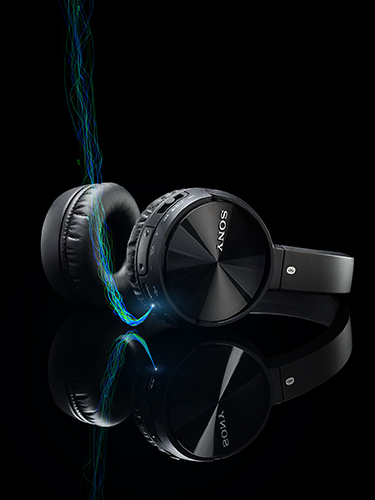 Black over-ear wireless headphones with a sleek design are positioned on a reflective black surface. A stream of multicolored light trails around them, creating a dynamic and futuristic effect.