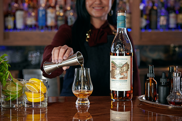 A bartender pouring whiskey from a jigger into a glass. A bottle of whiskey, fresh lemon and lime slices, mint, and small bottles of bitters are on the counter. Shelves of liquor bottles are blurred in the background.