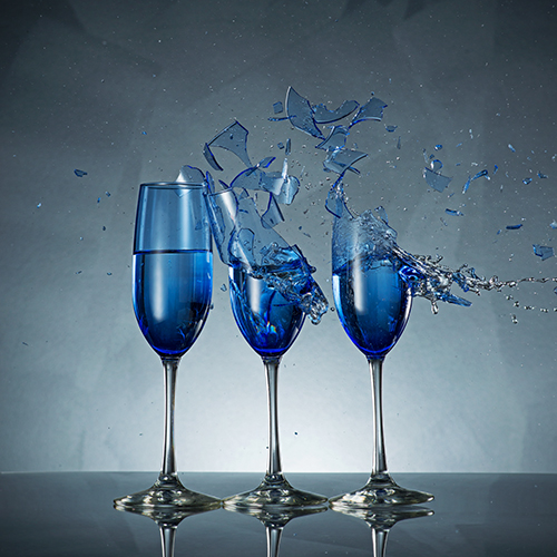 Three blue champagne flutes filled with liquid on a reflective surface. The flute on the right is shattering, with glass fragments and liquid splashing outward against a gray background.