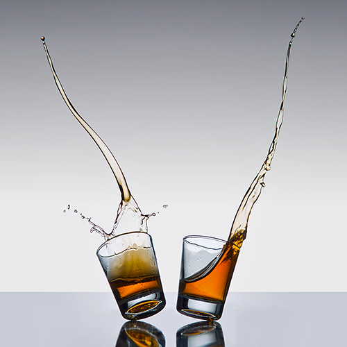 Two shot glasses of amber liquid are in motion, tilting away from each other on a reflective surface. Liquid splashes dramatically upward from both glasses against a gradient gray background.