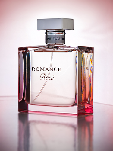 A bottle of Ralph Lauren Romance Rosé perfume is shown. The rectangular bottle features a gradient pink hue, with a silver cap reflecting light. It rests on a shiny surface against a softly lit background.