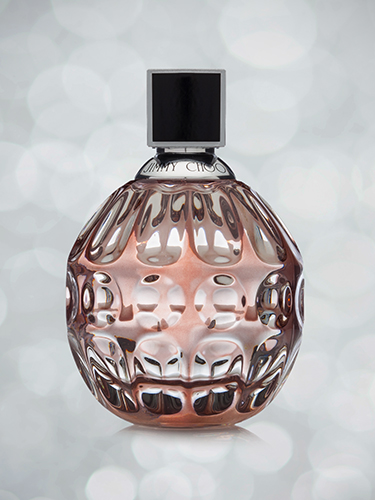 A Jimmy Choo perfume bottle with an intricate, textured design. It has a spherical shape with a metallic cap. The glass is tinted with a warm pink hue. The background is softly blurred with circular light patterns.