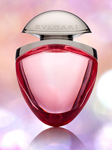 A bottle of BVLGARI perfume with a sleek, curved design and pink-tinted liquid inside. The bottle is topped with a silver cap, set against a blurred background of soft pastel colors