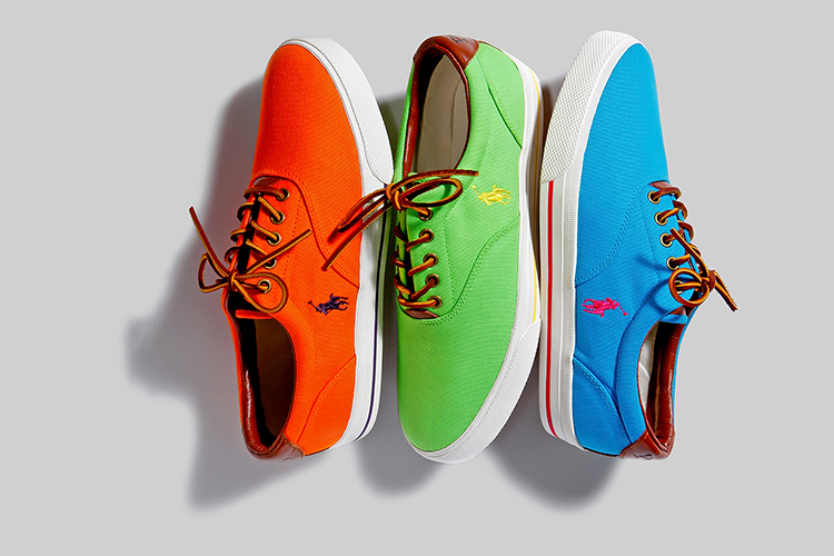 Three colorful sneakers are shown against a light gray background. The sneakers are orange, green, and blue, each featuring logos in contrasting colors and brown laces. They are arranged side by side, with shadows cast below.