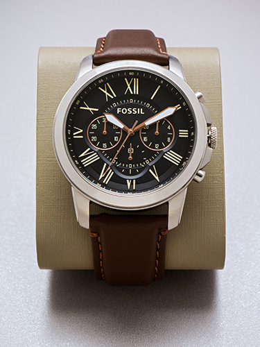 A Fossil watch with a brown leather strap is displayed on a stand. The watch face is black with Roman numerals and three smaller dials. The silver case contrasts with the brown strap and beige background.
