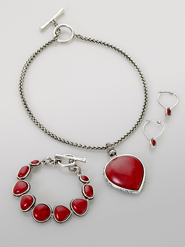 A jewelry set featuring a silver chain necklace with a large red heart-shaped pendant, a matching bracelet with red oval stones, and a pair of hoop earrings with red teardrop-shaped stones, all set against a plain gray background.