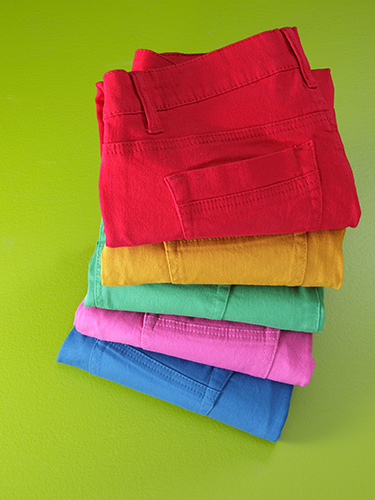 A stack of folded pants in vibrant colors: red, yellow, green, pink, and blue. The pants are neatly arranged on a bright green background.