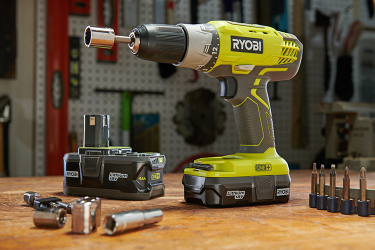Close-up of a Ryobi cordless drill on a wooden table, surrounded by a lithium battery, several drill bits, and sockets. A well-lit workshop background with various tools hanging on a perforated board