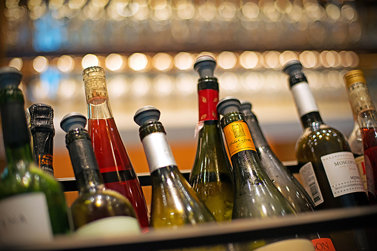 A collection of wine bottles in a rack, each with a variety of colored tops and labels, against a blurred background of glassware on shelves.