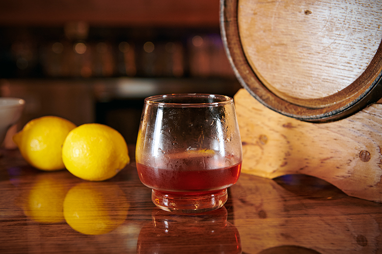 A glass of amber colored cocktail sits on a wooden bar counter, accompanied by two lemons and a wooden barrel in the background. The setting has a warm, inviting ambiance