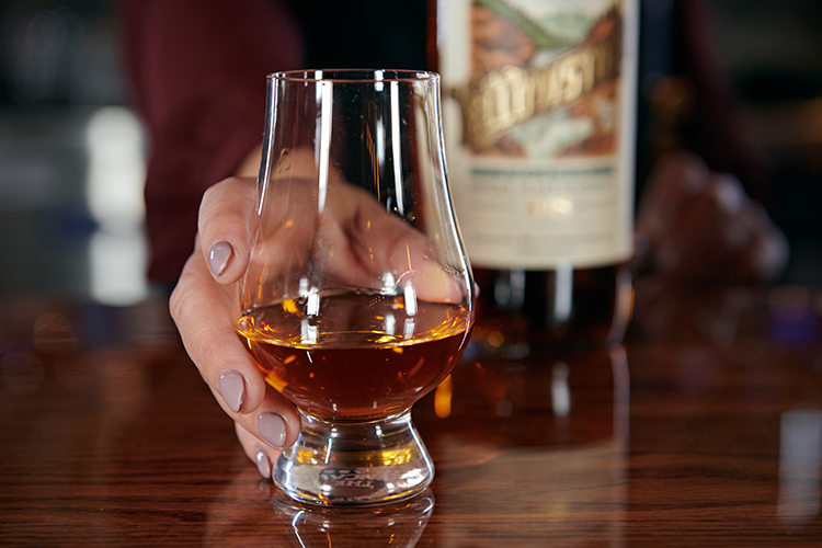 A hand with painted nails holds a glass of amber whiskey on a polished wooden table, with a blurred whiskey bottle in the background.