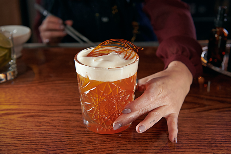 Close-up of a bartender presenting a cocktail in a textured glass they just mixed. The drink has a creamy foam top and a decorative caramel garnish.