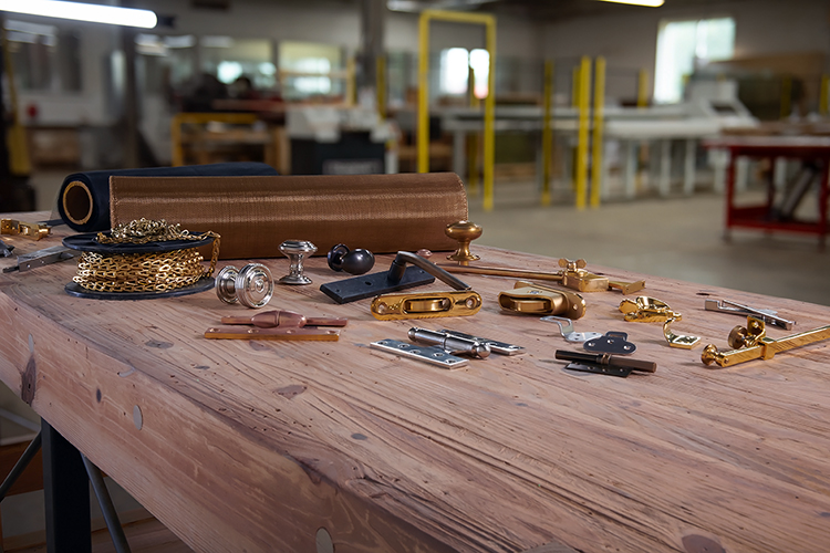 A wooden workbench displaying various hardware items, including brass and metal hinges, knobs, and fittings. In the background, a workshop with tools and equipment is partially visible.