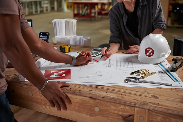Two people working at a table with a blueprint, a smartphone, a tape measure, a calculator, and a white helmet with a red logo. One is writing, and the other is leaning over the table.