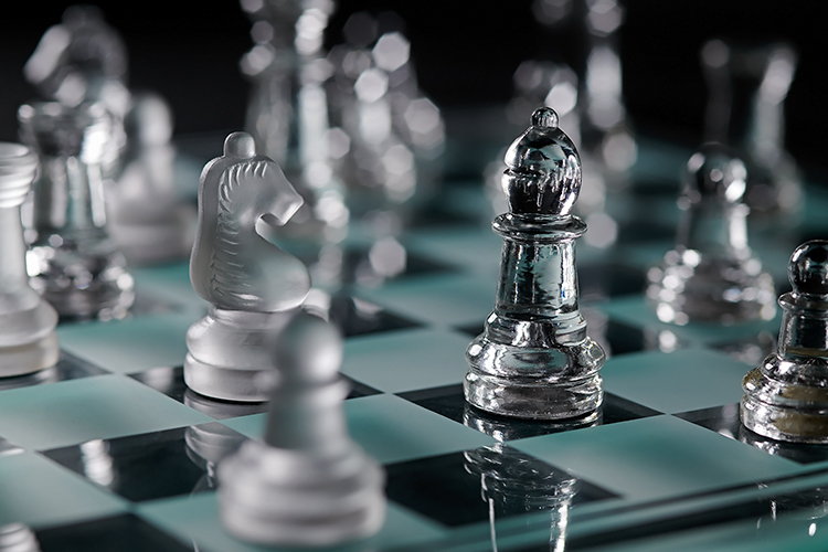 Close-up of a glass chessboard with transparent and frosted pieces. A frosted knight and a transparent bishop are prominently featured, surrounded by blurred pieces in the background, creating a reflective and strategic ambiance.